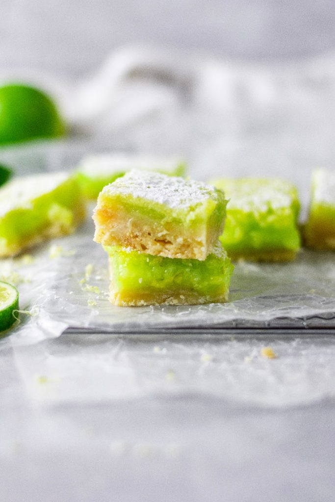 Two lime bars stacked on top of each other. They're sitting on a piece of wax paper with lime zest all around them.
