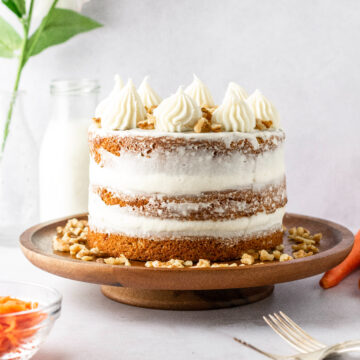 A semi-naked frosted carrot cake with milk in the background and carrots.