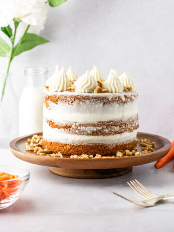 A semi-naked frosted carrot cake with milk in the background and carrots.