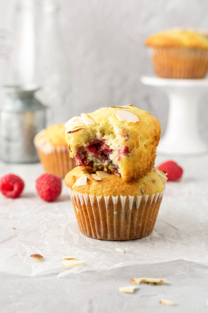 Raspberry muffins stacked on top of eachother with fresh raspberries in the background.