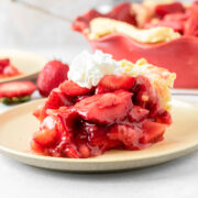 Strawberry pie slice with whipped cream and a red ceramic pie plate.
