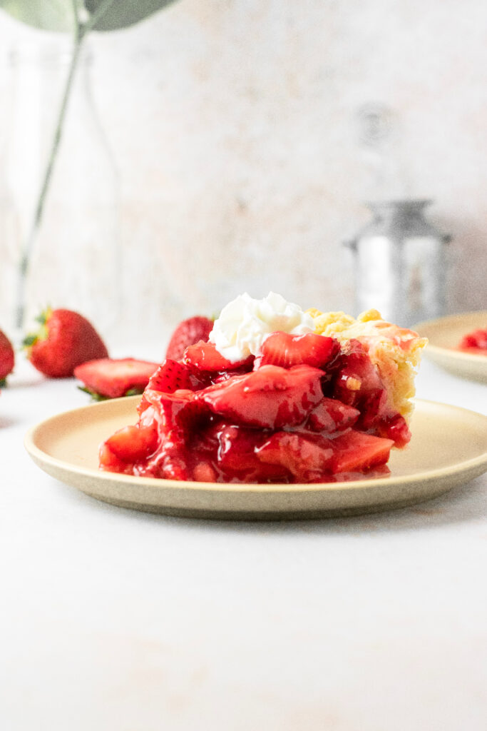A piece of strawberry pie sitting on a plate with a plant/vase in the background.