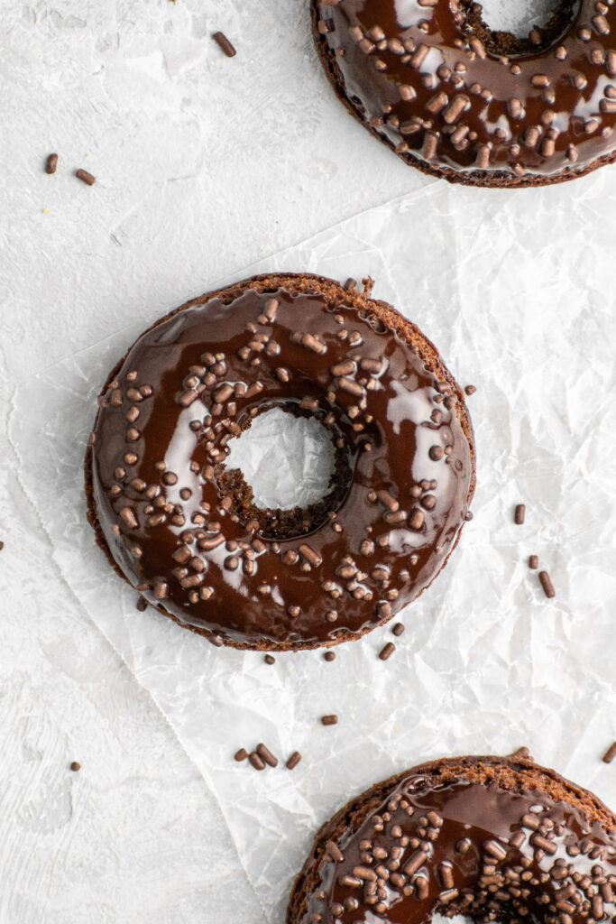 Double chocolate baked donuts sitting on parchment paper with chocolate sprinkles on the side.