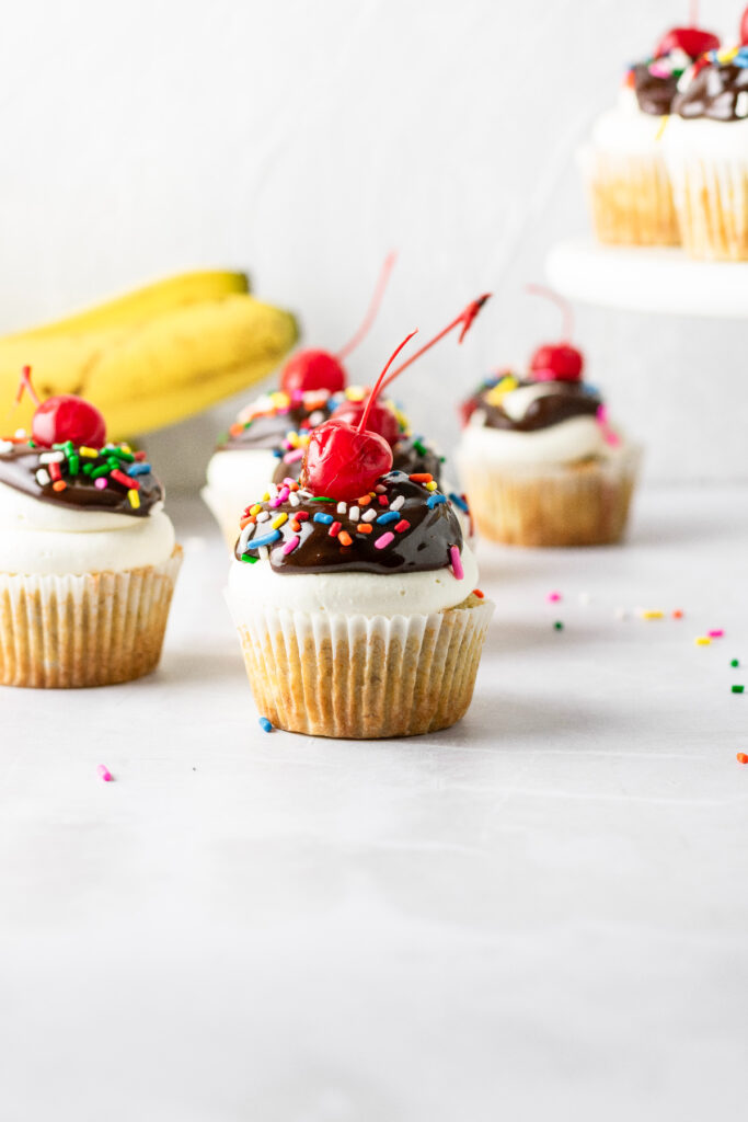 Banana split cupcakes with bananas in the background and sprinkles on the table.