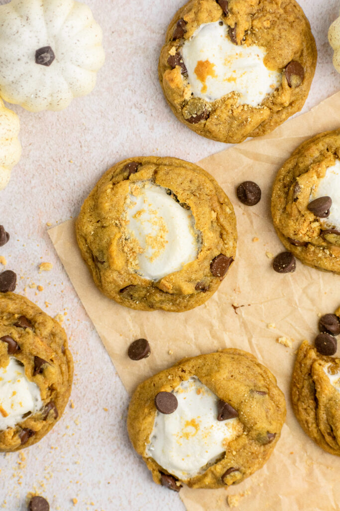 Pumpkin s'mores cookies on a brown parchment paper surrounded by chocolate chips.