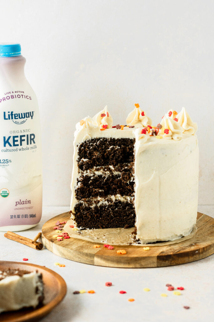 Chocolate cake with cream chese frosting sitting on a brown cake stand and a bottle of milk in the corner.