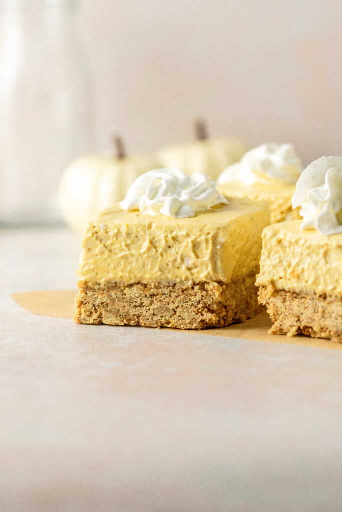 No bake pumpkin cheesecake bars with whipped cream on top and white pumpkins in the background.