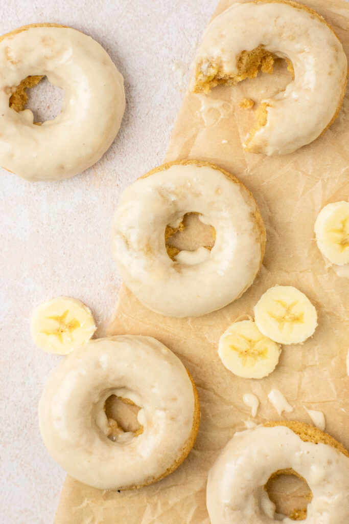 Banana glazed donuts sitting on top of parchment paper with sliced bananas on the side.