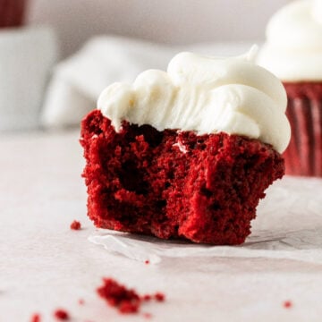 A red velvet cupcake topped with creamy cream cheese frosting with a bite taken out of it.
