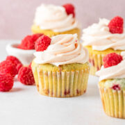 Four raspberry frosted cupcakes with a bowl on raspberries on the side.