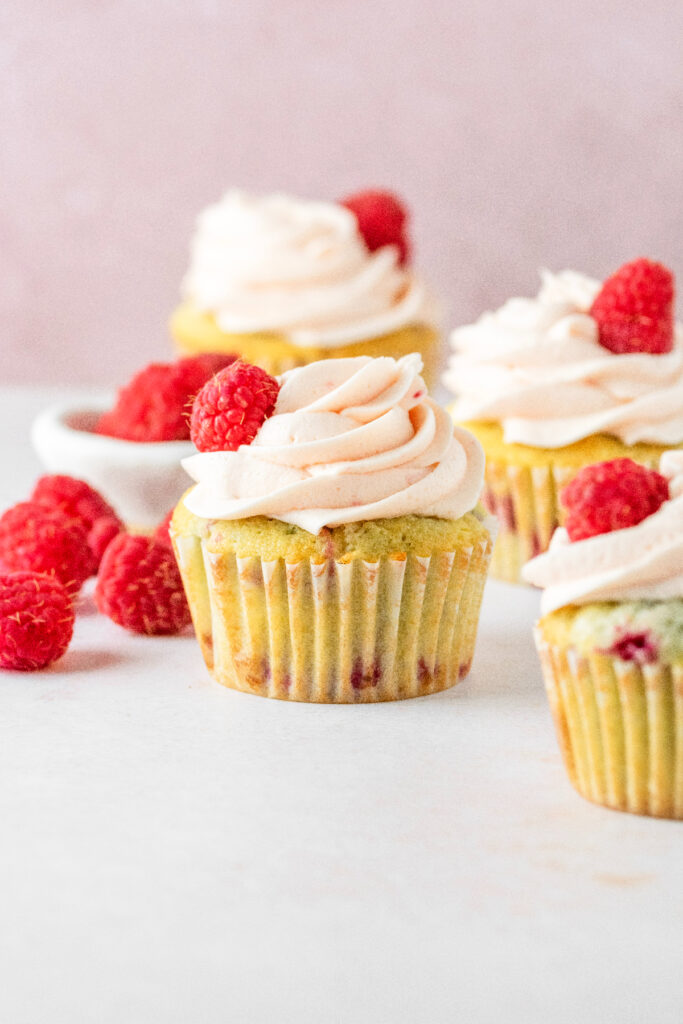 Four raspberry frosted cupcakes with a bowl on raspberries on the side.