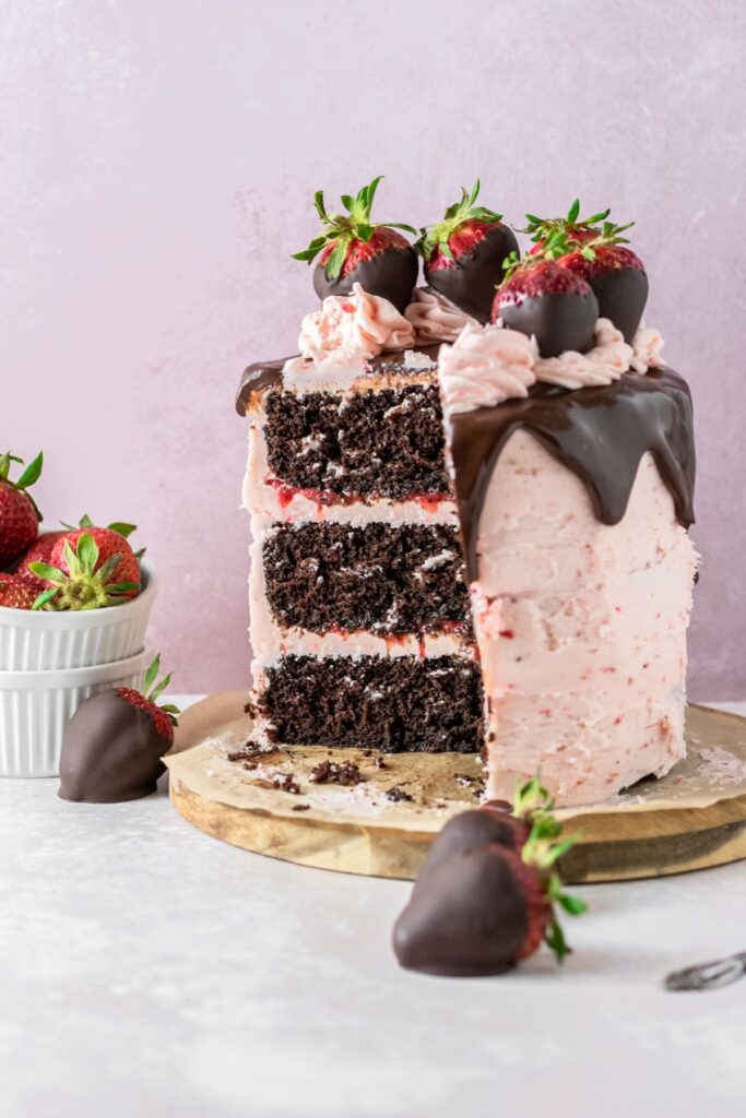 Chocolate layer cake with strawberry frosting and chocolate strawberries on top. It's sitting on a round brown cake board.