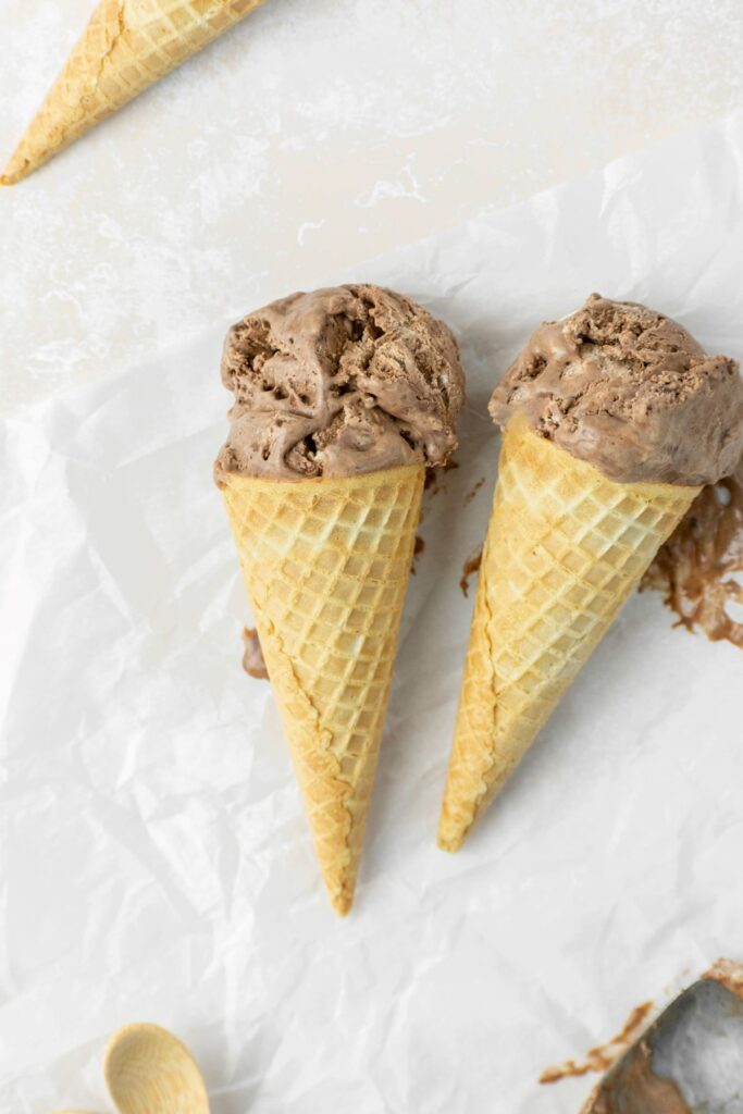 Two ice cream cones with chocolate ice cream and a wooden spoon on the side.