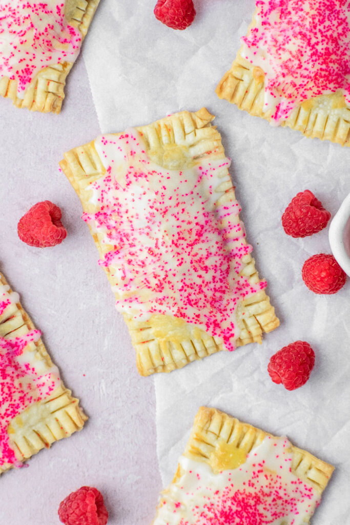 Raspberry toaster pastries with glaze and sprinkles on top. Fresh raspberries are scattered around them.