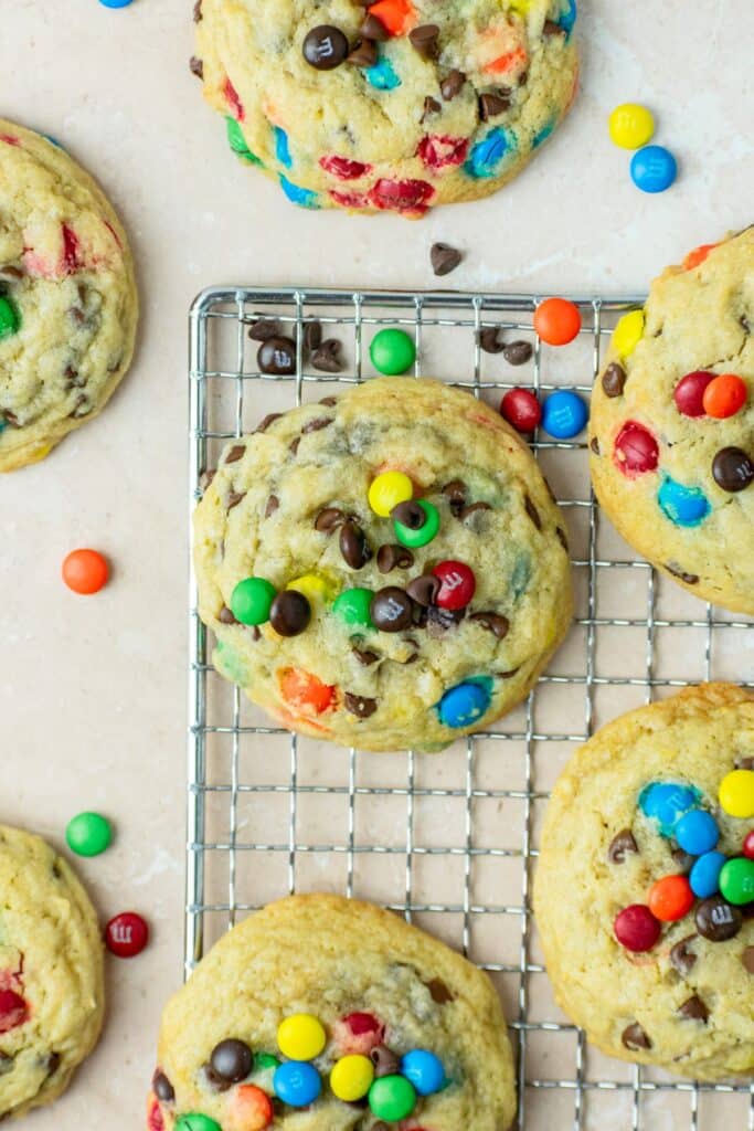 Many chocolate chip cookies with m&ms in them sitting on a wire rack.