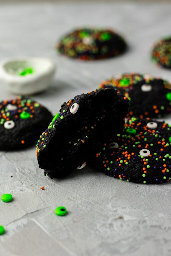 A Halloween cookie split in half. It's a pitch black color that has been rolled in green, purple, and orange sprinkles.