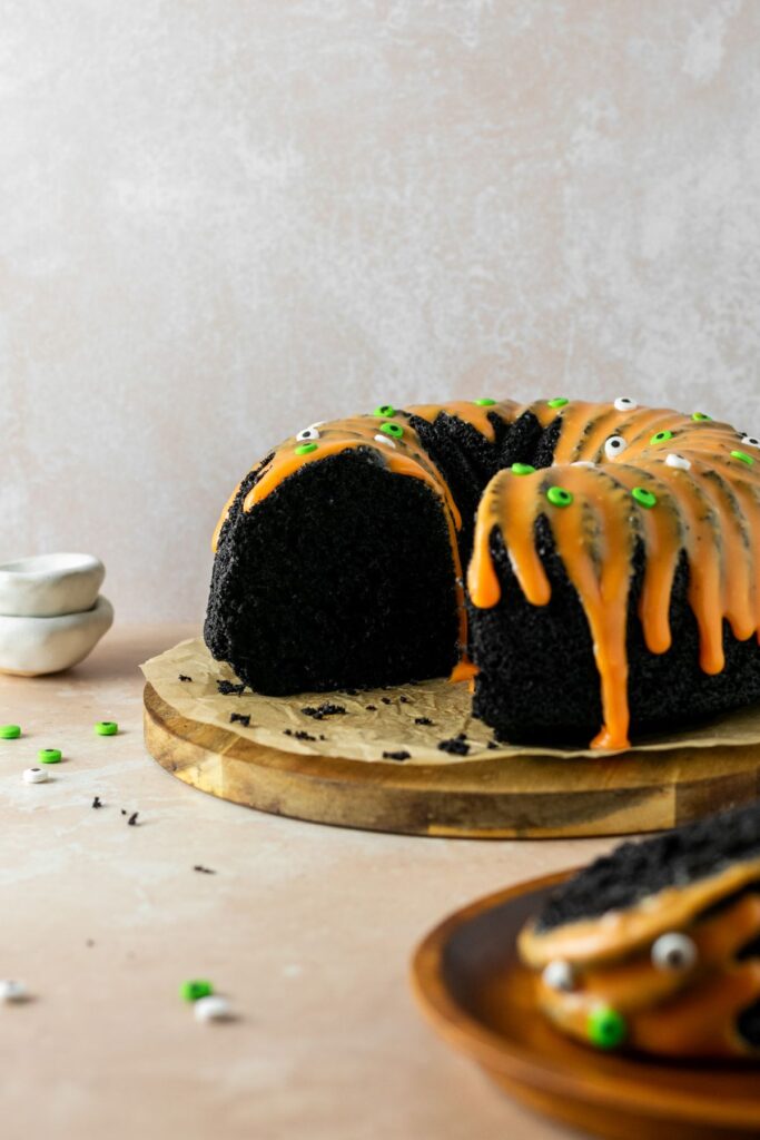 A black chocolate cake with orange icing on top. It's sitting on a wooden cake plate with green and white eyeball sprinkles.
