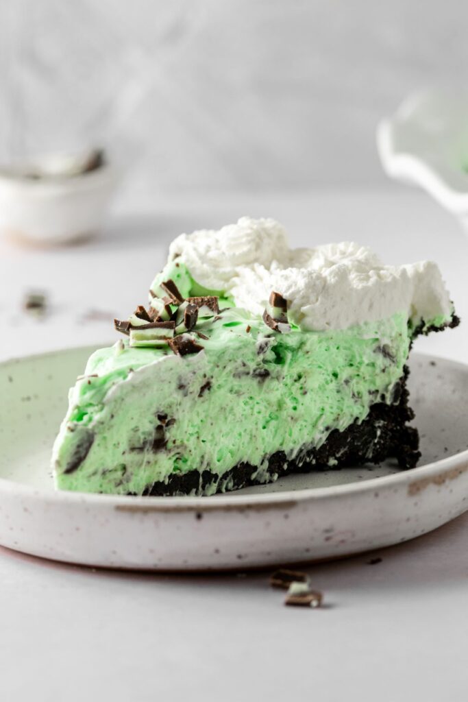 Andes mint pie slice on a white plate covered in extra candy pieces and homemade whipped cream.