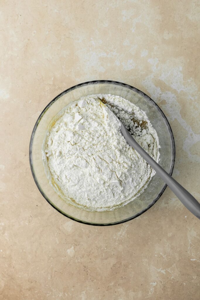 Flour added to wet ingredients in a large bowl with a rubber spatula.