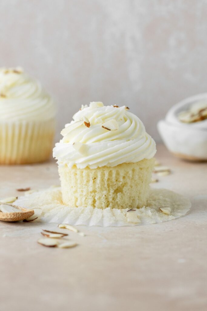 An almond cupcake with the cupcake liner peeled off of it. Almond slices are surrounding the cupake.
