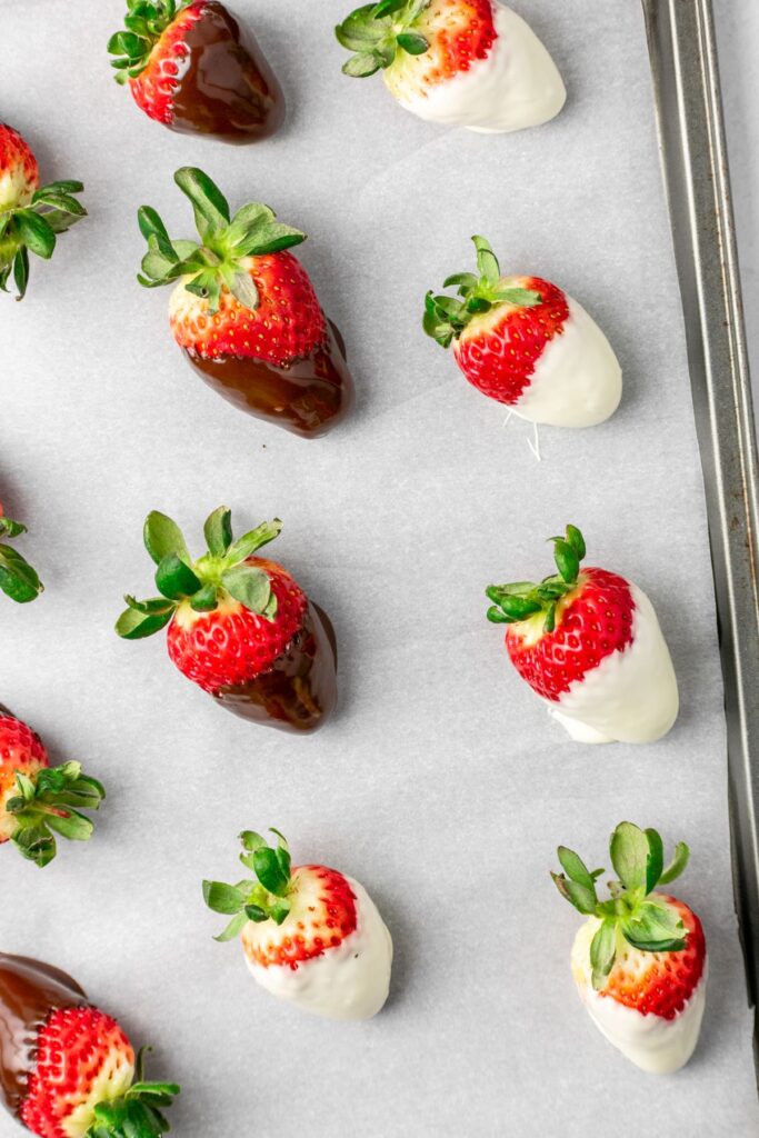 Strawberries on a parchment paper lined baking sheet that have been dipped in melted dark chocolate and white chocolate.