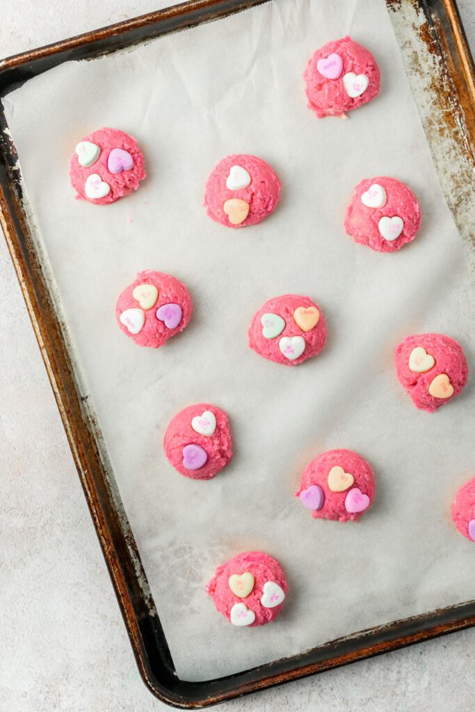 Pink cookie dough balls with heart candies pressed into them on a parchment paper lined baking sheet.