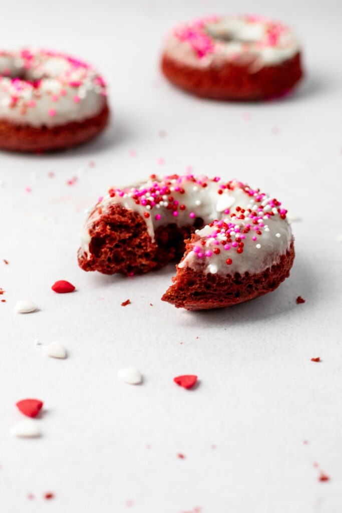 A bite taken out of a red velvet doughnut with heart sprinkles all around it.