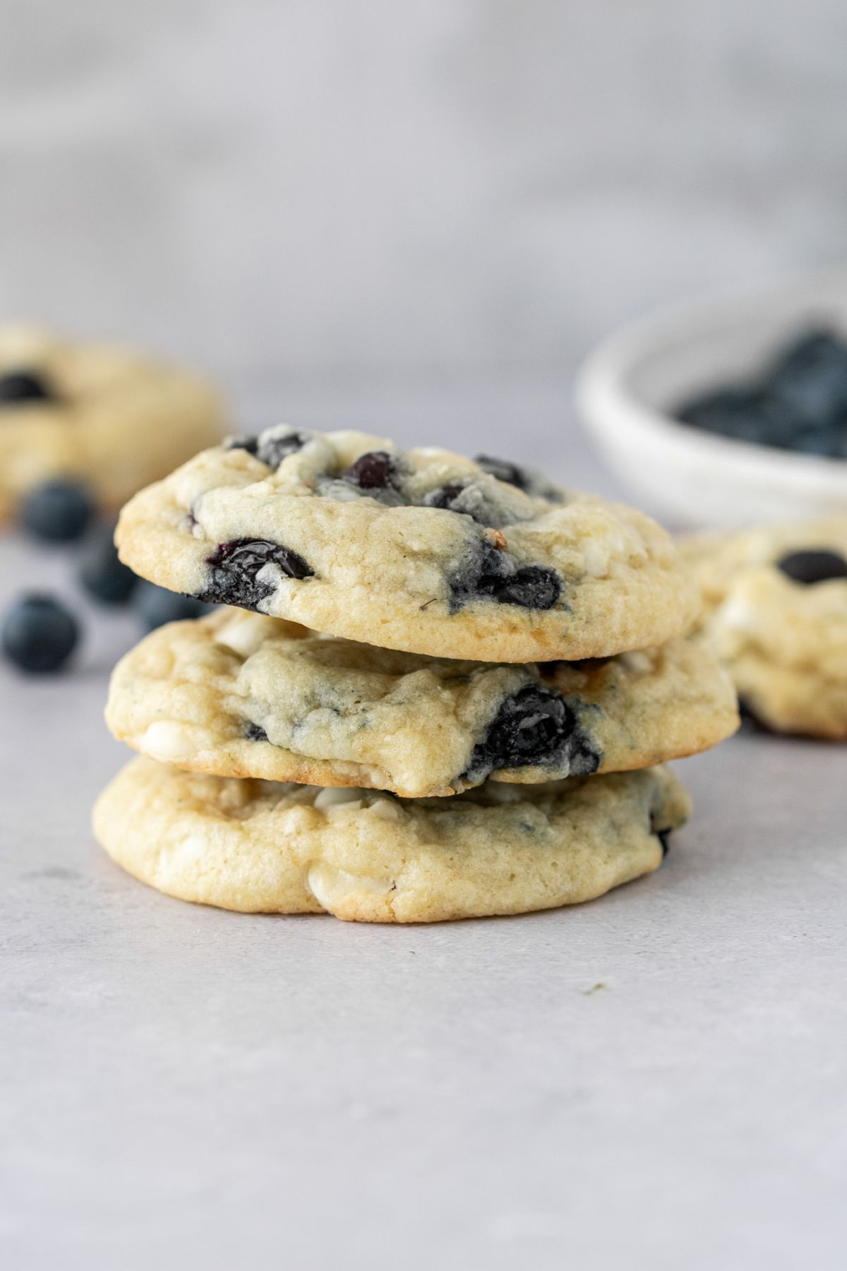 Three blueberry cookies stacked on top of each other with a plate of blueberries behind them.