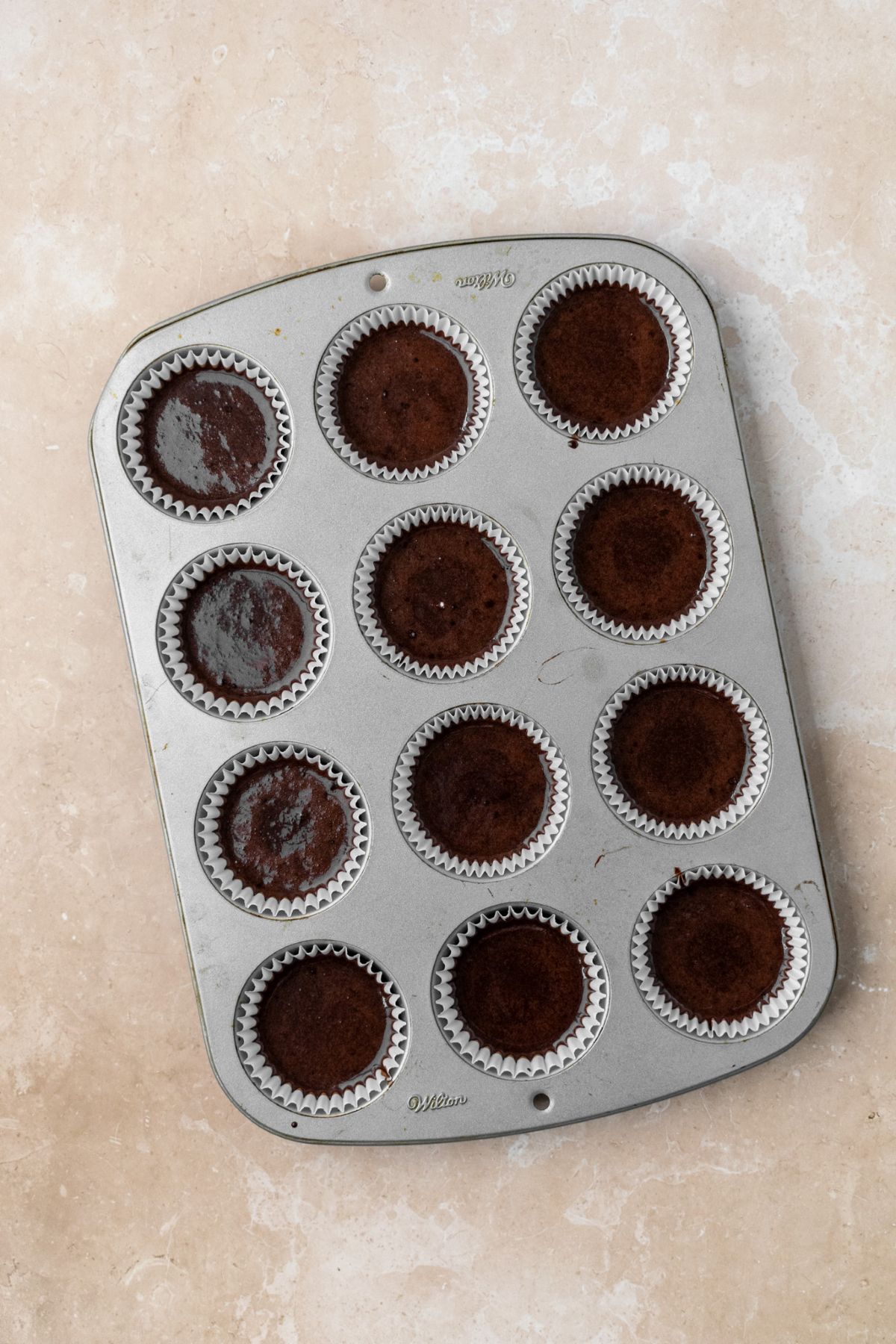 A muffin pan with white cupcake liners filled with chocolate cake batter.